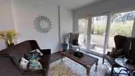 This thoughtfully designed single-floor space offers a seamless blend of comfort and style, centered around a well-appointed living area that serves as the heart of the home. The sitting room is finished with smooth white walls and clean white trim, creating a crisp, airy backdrop that enhances the sense of openness throughout. Durable wood-look flooring runs underfoot, adding warmth and texture to the refined interior.

A standout feature of the living area is the set of full-height glass French doors that open directly onto an outdoor balcony, flooding the room with generous natural light and framing lush greenery and surrounding architecture beyond. The seamless indoor-outdoor connection creates an inviting, resort-like atmosphere.

Decorative touches elevate the space with character — a sunburst-style wall mirror adds an artistic focal point, while a woven rattan coffee table, a classic wingback chair, and a plush area rug with a floral motif contribute layers of texture and elegance. The overall ambiance is calm and curated, striking a balance between casual comfort and refined aesthetic sensibility, making this space both visually appealing and highly functional for everyday living.