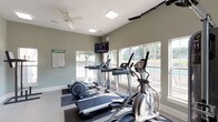 This fitness center offers a bright, well-organized space designed to accommodate a full range of workout routines. The interior is finished with neutral sage-green walls, light-toned flooring, and flush ceiling lighting panels that keep the environment evenly illuminated throughout the day. A ceiling fan aids in air circulation, contributing to a comfortable exercise atmosphere.

The cardio zone is positioned along a wall of large windows that frame views of an outdoor pool and surrounding greenery, allowing natural light to flood the space while providing a pleasant backdrop during workouts. Multiple treadmills and elliptical machines are arranged in this area, along with a wall-mounted flat-screen television for entertainment. Workout reference posters, including lower body and stretching guides, are displayed throughout for user convenience.

The strength training section houses a variety of cable and resistance machines, including a leg press and multi-function cable stations, providing options for both upper and lower body conditioning. Exercise mats and additional accessories are available for stretching and floor-based workouts. Full-length mirrors along one wall visually expand the space and assist with form during exercise. Glass-panel double doors lead to an adjoining area, enhancing the openness and accessibility of the facility. Overall, the fitness center delivers a clean, functional, and inviting environment for daily exercise.
