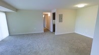 This single-floor space presents a clean, functional layout designed for comfortable living. The main living area is generously sized, featuring wall-to-wall carpeting in a neutral beige tone that adds warmth underfoot while complementing the soft, light green walls and crisp white ceiling. The open feel of the room is enhanced by its simple, uncluttered design and smooth white baseboards that frame the space neatly.

A central hallway extends from the main living area, leading to additional rooms within the unit. Wall-mounted light fixtures line the hallway, providing consistent illumination throughout. Partially visible from the main space is what appears to be a kitchen area, identifiable by dark cabinetry, offering a practical contrast to the lighter tones found throughout the rest of the space.

An electrical panel is mounted on the wall near the hallway entrance, a functional detail that is cleanly integrated into the overall aesthetic. The neutral color palette of beige, white, and soft green is carried consistently throughout, creating a cohesive and calm ambiance. Overall, the space offers a straightforward, move-in-ready environment with a layout that supports easy furniture arrangement and everyday functionality.