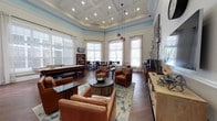 Located at 2222 Crow Ln in Myrtle Beach, SC, this single-floor community clubroom is a well-appointed gathering space designed for both relaxation and recreation. The room immediately impresses with its soaring coffered ceiling, accented by a soft blue crown molding band and recessed lighting, lending an air of elegance and spaciousness throughout.

Rich hardwood flooring runs the length of the space, complementing the warm tones of the leather club chairs and the deep wood finishes of the furniture. A full-size pool table serves as a central recreational feature, positioned beneath stylish pendant lights that add a warm, inviting glow. Adjacent to the game area, a comfortable lounge zone features a grouping of cognac leather armchairs arranged around a dark round coffee table, anchored by a teal-accented area rug, with a large flat-screen TV mounted nearby for entertainment.

A dedicated café-style dining area lines one wall, furnished with dark high-top tables and bar stools, all bathed in natural light streaming through the numerous floor-to-ceiling windows dressed with sheer curtains and blinds. A bar counter with dark granite surfaces and rustic wood stools provides a convenient refreshment station, complete with a coffee setup. Decorative wall art, sconce lighting, and carefully chosen accessories complete the polished yet welcoming aesthetic of this versatile amenity space.