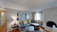 This single-floor space showcases a thoughtfully designed living area that balances comfort and contemporary style. The living room serves as the focal point, featuring a curved sectional sofa in soft blue-grey upholstery accented with mustard yellow throw pillows, adding a warm contrast to the cool color palette. A matching ottoman and accent chair round out the seating arrangement, all positioned atop a plush grey area rug that anchors the space over the hardwood flooring beneath.

Natural light filters into the room through a window dressed with sheer grey curtains paired with horizontal blinds, providing both brightness and privacy. The walls are finished in a neutral grey-beige tone, complemented by a textured white brick accent wall that adds architectural interest. Two matching decorative wall art pieces featuring architectural motifs hang prominently, contributing to the refined aesthetic.

A sleek side table with a geometric base supports a classic table lamp, offering warm ambient lighting. The overall ambiance is calm and cohesive, with a neutral foundation punctuated by carefully chosen accents that give the space a modern, inviting character.