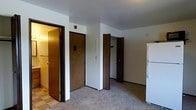 This single-floor residential unit offers a straightforward and functional layout designed to maximize usability. The main living area is finished with wall-to-wall carpet, providing a comfortable underfoot feel throughout the space. White walls keep the interior bright and neutral, complementing the textured ceiling overhead.

A notable feature of this unit is its storage capacity, with multiple closets accessible via dark wood-toned doors. One closet is equipped with a hanging rod, making it well-suited for use as a wardrobe. An additional door leads to a secondary room or utility area, adding to the overall versatility of the floor plan.

The entryway is defined by a solid wood-toned door fitted with a standard lock, alongside a fire alarm mounted at the top — indicating compliance with safety standards. A wall-mounted panel, likely for lighting or intercom controls, is conveniently positioned near the entry.

The kitchen area is partially visible and includes a full-size white refrigerator, suggesting a practical kitchen setup. The flooring transitions from carpet to a tiled surface in this zone, clearly delineating the living and kitchen spaces. Overall, the unit presents a clean, move-in-ready environment suited for everyday residential living.