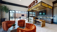 Located at 1333 N Santa Fe Ave in Vista, California, this single-floor social lounge is a thoughtfully designed gathering space that blends industrial charm with upscale comfort. The centerpiece of the space is a fully equipped bar area, crowned by a bold marquee "CHEERS" sign, featuring dark stone countertops, a built-in draft beer tap, under-counter refrigeration, a decorative bottle-cap display wall, and a warm marble-toned backsplash — all framed in rich wood cabinetry and black metal accents. A large garage-style door opens the bar directly to an outdoor patio, seamlessly extending the social experience to the exterior. The lounge area is anchored by a striking exposed brick accent wall and patterned tile flooring, setting a warm, eclectic tone. Plush tufted velvet armchairs in deep burnt orange, a tufted leather sofa, and a live-edge wood coffee table create an inviting conversational seating area beneath an industrial pendant light. A secondary lounge zone features high-top tables and bar stools positioned along large windows that flood the space with natural light and offer views of the surrounding neighborhood. Dark hardwood flooring, teal velvet seating, and layered area rugs complete the vibrant, curated aesthetic throughout.