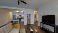 This single-floor residence offers a thoughtfully designed open-concept layout that integrates the living, dining, and kitchen areas into one cohesive and functional space. The living area features rich, dark hardwood-style flooring that flows throughout, anchored by a classic ceiling fan that enhances comfort and airflow. A wooden coffee table and plush seating create a welcoming focal point, while an open metal-and-wood shelving unit adds a rustic-industrial accent between spaces. The kitchen is well-equipped with stainless steel appliances, including a range, microwave, and refrigerator, complemented by crisp white cabinetry, recessed lighting, and a peninsula with bar seating — ideal for casual dining or entertaining. Pendant lighting adds a stylish touch above the counter. The space is finished in neutral tones throughout, with clean white walls that amplify the sense of openness and brightness. A hallway visible from the main living area leads to additional rooms, suggesting a practical and well-organized floor plan. Overall, the space strikes a balance between modern functionality and warm, inviting aesthetics.