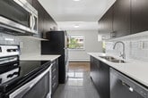 Located at 2067-2077 Prospect St in Burlington, Ontario, this one-bedroom, two-bathroom unit offers a well-designed single-floor layout that makes efficient use of its space. Throughout the living areas and bedroom, warm-toned parquet hardwood flooring adds a classic character that contrasts nicely with the clean, neutral-painted walls and white trim.

The combined kitchen and living area forms the heart of the unit. The galley-style kitchen is outfitted with rich dark wood cabinetry, white countertops, a tile backsplash, stainless steel appliances including a full-size refrigerator, and polished tile flooring — providing both style and functionality. The adjacent living room is bright and open, with a sliding glass door that opens to a private balcony overlooking a landscaped outdoor area with views of neighbouring greenery.

The bedroom is a comfortable, well-proportioned room featuring a window that allows natural light to fill the space, a baseboard heating unit, and a built-in closet with bi-fold doors for ample storage. Two bathrooms serve the unit, one of which features warm taupe subway tile surrounding a soaker tub and shower combination, paired with a dark wood vanity and clean white countertop.

A hallway connects the rooms and contributes to the unit's practical flow. Overall, the space presents a blend of classic and contemporary finishes in a residential Burlington location.