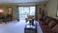 This single-floor space centers around a well-appointed living room that exudes a warm and inviting ambiance. Large sliding glass doors dominate one wall, flooding the room with natural light and providing direct access to a private balcony overlooking a landscaped outdoor setting with mature trees. The generous expanse of windows also frames pleasant views of the surrounding greenery, enhancing the sense of openness throughout the space.

The living room is thoughtfully arranged with comfortable seating, including a plush armchair positioned near the sliding doors and a rich burgundy sofa anchoring one side of the room. A dark-finished coffee table serves as a central focal point, elegantly styled with decorative vases and a metallic tray. Neutral-toned carpeting runs throughout, complementing the warm earth tones of the furnishings.

Decorative accents, including a tall sculptural vase, wall-mounted ornamental plates, and lush potted plants, add character and depth to the room. Curtain rods with rust-colored drapes frame the windows, contributing to the cohesive, earthy color palette. The overall atmosphere is comfortable, stylish, and filled with natural light.
