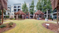Located at 415 Armour Dr in Atlanta's vibrant 30324 zip code, this thoughtfully designed outdoor courtyard serves as a central gathering space for its multi-story residential community. The courtyard is anchored by two striking wooden pergolas with decorative lattice detailing, positioned symmetrically to frame a well-maintained open lawn area. Their warm wood tones complement the classic brick-and-neutral-stucco facade of the surrounding building, which features multiple floors of private balconies overlooking the grounds below.

Winding brick-paver pathways connect the various areas of the courtyard, guiding residents through carefully curated landscaping that includes mature shade trees, manicured shrubs, and mulched garden beds. A stone-ringed fire pit serves as a natural focal point in one corner of the space, accompanied by seating that invites outdoor gatherings. Bollard-style pathway lighting is integrated throughout, ensuring the courtyard remains functional and inviting into the evening hours.

The overall ambiance is one of serene, park-like retreat within an urban setting — a private green escape that balances open lawn space with shaded pergola areas and intimate garden nooks. The surrounding architecture enhances the sense of enclosure and community, making this courtyard a standout amenity of the property.
