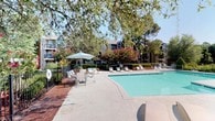 This single-story residence is part of a well-established residential community defined by its classic brick exterior and thoughtfully maintained grounds. The building's exterior showcases traditional red brick construction accented by light-colored lap siding trim, with large sliding glass windows fitted with horizontal blinds that allow for natural light and outdoor views. A private patio area sits just outside the unit, bordered by neatly shaped brick planters filled with lush greenery, creating a sense of intimate outdoor space.

The surrounding landscape is richly planted with mature trees, ornamental shrubs, and ground cover, offering a green and peaceful environment. Wrought iron fencing lines the perimeter walkways, framing views of mature oak trees and well-kept lawns that extend toward the street. Colorful flowering shrubs, including vibrant crepe myrtles, add seasonal color throughout the grounds.

A standout feature of the community is the expansive outdoor pool area, complete with a generously sized swimming pool, a broad concrete pool deck, and multiple lounging chairs and shaded seating arrangements. Mature trees and lush flowering shrubs surround the pool, creating a private, resort-like atmosphere ideal for relaxation. The overall ambiance of this community is one of comfort, greenery, and classic residential character.