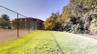 This outdoor dog park offers a well-maintained, fully enclosed space designed for pets to roam freely in a safe environment. The perimeter is secured by sturdy chain-link fencing, providing clear boundaries while maintaining open sightlines to the surrounding landscape. The grassy area is generously sized, offering ample room for exercise and play beneath open skies, with mature trees lining the backdrop and providing natural shade along the edges.

Adjacent to the dog park is a hard-surface tennis court, also enclosed by chain-link fencing, adding to the overall recreational amenities of the property. The courts benefit from full sun exposure and are set against a backdrop of mature trees and a multi-story brick building nearby.

A covered entry pavilion constructed of classic red brick serves as the access point to the facility. The pavilion features a concrete floor, a wall-mounted intercom system with an informational notice board, and a glass-paneled entry door with a keypad access panel. Electrical outlets are integrated into the brick walls, and the covered overhang provides shelter at the entry point. The overall setting is open, functional, and community-oriented.