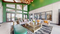 Located at Tomoka Town Center in Daytona Beach, Florida, this single-floor community clubhouse offers a generously sized, thoughtfully designed gathering space that blends contemporary style with inviting comfort. The space is divided into two distinct living areas, each with its own personality and purpose.

The first living area immediately captures attention with its dramatic double-height ceilings clad in warm wood paneling, bold emerald green accent walls, and a striking sculptural chandelier composed of clustered globe lights. Floor-to-ceiling windows along the perimeter flood the space with natural light and offer open views of the surrounding exterior. Large-format tile flooring and an array of stylishly arranged seating — including plush sofas, accent chairs, and a teal leather bench — create a welcoming and visually dynamic atmosphere.

The second living area offers a more relaxed, sophisticated ambiance with a coffered ceiling detailed with wooden beams, a textured mosaic accent wall, and large windows draped with warm linen curtains. Light wood-look tile flooring extends throughout, anchoring a curated seating arrangement designed for conversation and lounging.

A dedicated game room completes the space, featuring a full-sized billiards table, a large-format wall-mounted television, a marble-topped kitchen island with bar seating, teal subway tile backsplash, white cabinetry, and eye-catching angled wooden ceiling slats — all accessible through expansive garage-style glass doors that open to the outdoors.