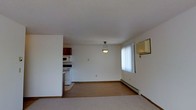This single-floor space offers a practical, open-concept layout where the living area and kitchen flow seamlessly into one another. The interior is finished with white walls and a textured ceiling, creating a clean, neutral backdrop throughout. The living area is carpeted, providing a comfortable underfoot feel, while the kitchen transition introduces a hard flooring surface. A pendant light fixture hangs centrally, serving as a modest focal point in the open space. Natural light enters through a window fitted with vertical blinds, brightening the area. A baseboard heater runs along the exterior wall beneath the window, indicating a reliable heating source. The kitchen is positioned to one side and features dark wood cabinetry, a white electric range, and a built-in microwave, offering functional cooking space. The layout is straightforward and efficient, making good use of the available floor space. Overall, the space presents a simple, utilitarian design with a light and airy feel suited to everyday living.