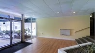 This single-floor commercial space offers a clean, functional layout designed to accommodate a variety of business uses. The main area features warm wood-look flooring that contrasts neatly with soft yellow-toned walls, creating a welcoming and professional atmosphere. A drop ceiling with a grid tile system houses recessed lighting throughout, ensuring consistent, even illumination across the space.

A prominent floor-to-ceiling glass entrance and large window panel along one wall floods the interior with natural light and provides clear views of the surrounding wooded landscape outside. This glass frontage also creates a strong visual connection between the interior and exterior, enhancing the sense of openness. A dark entry mat at the threshold provides a practical transition from the outdoors.

A wall-mounted HVAC unit is positioned along one of the interior walls, offering climate control directly within the main area. The space is otherwise open and unobstructed, providing flexibility for office, retail, or service-oriented configurations. The clean, neutral finishes and well-maintained surfaces throughout reflect a move-in-ready condition, making this a practical and adaptable commercial environment suited to a wide range of professional tenants or owner-occupants.