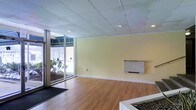 This single-floor commercial space offers a clean, functional layout designed to accommodate a variety of business uses. The main area features warm wood-look flooring that contrasts neatly with soft yellow-toned walls, creating a welcoming and professional atmosphere. A drop ceiling with a grid tile system houses recessed lighting throughout, ensuring consistent, even illumination across the space.

A prominent floor-to-ceiling glass entrance and large window panel along one wall floods the interior with natural light and provides clear views of the surrounding wooded landscape outside. This glass frontage also creates a strong visual connection between the interior and exterior, enhancing the sense of openness. A dark entry mat at the threshold provides a practical transition from the outdoors.

A wall-mounted HVAC unit is positioned along one of the interior walls, offering climate control directly within the main area. The space is otherwise open and unobstructed, providing flexibility for office, retail, or service-oriented configurations. The clean, neutral finishes and well-maintained surfaces throughout reflect a move-in-ready condition, making this a practical and adaptable commercial environment suited to a wide range of professional tenants or owner-occupants.