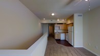 This single-floor residential space offers a practical and functional layout designed for comfortable everyday living. The open-concept design connects the living area to the kitchen, creating a sense of continuity throughout the unit. The living space is finished with wall-to-wall carpeting, adding warmth and a soft underfoot feel, while the kitchen transitions to a hard flooring surface for easy maintenance. The kitchen is well-equipped, featuring light wood cabinetry that provides ample storage, a double sink, and white appliances including a refrigerator, dishwasher, and range. A track lighting fixture illuminates the kitchen area, complementing the flush-mount ceiling light that brightens the main living space. The neutral, warm-toned walls throughout contribute to a cohesive and inviting ambiance. A hallway leads to a bathroom, visible in the background, offering a straightforward and efficient floor plan. The layout maximizes the use of available space, making it a well-organized and accessible living environment suitable for a range of occupants.