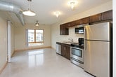 This single-floor unit showcases a contemporary industrial aesthetic, characterized by polished concrete floors, exposed metallic ductwork, and light wood trim accents that run throughout the space. The open-concept kitchen is outfitted with dark espresso cabinetry, stainless steel appliances including a refrigerator, range, microwave, and dishwasher, complemented by a clean countertop with an undermount sink. Industrial-style flush-mount light fixtures add to the urban character of the kitchen and living areas.

The living area benefits from natural light through a wood-framed window offering views of surrounding brick architecture, with a ceiling fan providing additional comfort. A barn-style sliding door adds a modern rustic touch to the space. In-unit laundry is conveniently positioned near the entryway.

The unit includes two bathrooms: one featuring a dark-stained vanity with a framed mirror and tiled shower surround, and another with a floating dark vanity, also with a framed mirror and tiled shower. A dedicated bedroom with carpet flooring and a ceiling fan provides a quieter retreat. Additional storage is offered through a walk-in closet with wire shelving. The overall ambiance is modern, functional, and distinctly urban.