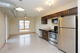 This single-floor unit showcases a contemporary industrial aesthetic, characterized by polished concrete floors, exposed metallic ductwork, and light wood trim accents that run throughout the space. The open-concept kitchen is outfitted with dark espresso cabinetry, stainless steel appliances including a refrigerator, range, microwave, and dishwasher, complemented by a clean countertop with an undermount sink. Industrial-style flush-mount light fixtures add to the urban character of the kitchen and living areas.

The living area benefits from natural light through a wood-framed window offering views of surrounding brick architecture, with a ceiling fan providing additional comfort. A barn-style sliding door adds a modern rustic touch to the space. In-unit laundry is conveniently positioned near the entryway.

The unit includes two bathrooms: one featuring a dark-stained vanity with a framed mirror and tiled shower surround, and another with a floating dark vanity, also with a framed mirror and tiled shower. A dedicated bedroom with carpet flooring and a ceiling fan provides a quieter retreat. Additional storage is offered through a walk-in closet with wire shelving. The overall ambiance is modern, functional, and distinctly urban.