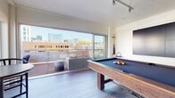 This contemporary amenity space is designed to offer a premium recreational and social experience across a single level. At its heart is a well-appointed billiards room, centered around a handsome wood-framed pool table with a slate-blue felt surface. Track lighting runs along the white ceiling, providing focused illumination throughout the space. One wall is fitted with a large multi-panel display screen, ideal for entertainment, while an adjacent area features a stylish wet bar with decorative tiled backsplash and built-in cabinetry. Light wood-look flooring runs throughout, contributing to the clean, modern aesthetic.

Floor-to-ceiling sliding glass doors line the exterior walls, flooding the interior with abundant natural light and providing seamless access to the outdoor terrace. The terrace itself extends generously along the building's exterior, clad in warm wood decking and equipped with comfortable lounge seating and bistro-style tables. A sleek linear fire pit serves as a focal point, making the outdoor space usable across seasons. The building's striking red paneled exterior facade adds a bold architectural element to the terrace setting.

Panoramic city skyline views are visible from both the interior and the terrace, creating an energetic urban backdrop. The overall ambiance is modern, vibrant, and ideally suited for both casual gatherings and social events.