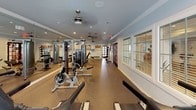 This fitness center is a comprehensively outfitted single-floor facility designed to support a full range of workout routines. The space is finished with durable, cork-style flooring throughout, providing a practical and comfortable surface for exercise. Crown molding and light blue-gray walls lend the room a polished, clean aesthetic that sets it apart from a typical gym environment.

The cardio zone features multiple treadmills with integrated display screens, an elliptical trainer, and a stationary bike, all positioned along a wall of large windows fitted with horizontal blinds. This arrangement allows for generous natural light while maintaining privacy. Ceiling fans are installed throughout, ensuring comfortable air circulation during workouts.

The strength training area is well-appointed with a multi-station cable machine, a dedicated weight bench, and a dumbbell rack offering a range of free weights. A full-length wall mirror expands the sense of space and allows users to monitor their form. A medicine ball storage rack and additional functional training equipment round out the offerings.

Access to the fitness center is provided through elegant dark-framed French doors on multiple sides of the room, with exterior views visible through the glass. Recessed lighting supplements the natural light, and wall-mounted artwork adds a welcoming touch to the overall environment.