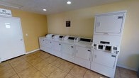 This single-floor laundry facility is designed for shared use, offering a practical and well-organized space for handling laundry needs. The room is outfitted with a row of top-loading washing machines aligned along the wall, complemented by a stacked washer-dryer unit, providing ample capacity for multiple users. The machines appear to be coin-operated or card-operated commercial-grade appliances, suitable for high-frequency use. The interior features warm yellow-painted walls that lend a bright and welcoming feel to the utilitarian space. Recessed ceiling lights provide even illumination throughout the room, while a textured ceiling adds a subtle decorative element. The flooring consists of durable, easy-to-clean beige ceramic tiles, well-suited for a high-traffic laundry environment. A posted laundry room rules and information sign is visible on the wall, indicating this is a shared-use facility. The layout is straightforward and efficient, maximizing the available floor space while keeping the machines easily accessible. Overall, the room is clean, functional, and well-maintained, providing a convenient on-site laundry solution.