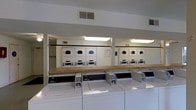 This shared laundry facility is a practical, single-floor space designed to accommodate the laundry needs of multiple users efficiently. The room is outfitted with a row of front-loading, coin-operated washing machines positioned along the main wall, providing ample capacity for simultaneous loads. Above and behind, stacked dryer units are neatly arranged, maximizing the use of vertical space and ensuring a high throughput for users. A continuous wooden folding counter runs along the top of the washing machines, offering a convenient surface for sorting and folding laundry. The room is well-lit with overhead fluorescent lighting, ensuring clear visibility throughout. A large mirror on one wall visually expands the space and enhances the sense of openness. Ventilation is provided through ceiling-mounted HVAC vents, maintaining a comfortable environment during use. The clean, white cabinetry housing the dryers and the neutral walls contribute to a bright, functional atmosphere. Overall, the space is thoughtfully laid out to support efficient laundry operations in a communal setting.