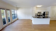 This single-floor residence offers a well-designed open-concept layout that seamlessly connects the living and kitchen areas into one cohesive, airy space. The living area is bathed in natural light streaming through large windows that frame views of lush greenery outside, creating a warm and inviting atmosphere. Smooth, light-toned hardwood flooring runs throughout, adding a clean and contemporary feel to the interior.

The kitchen is a standout feature, appointed with crisp white cabinetry that extends to the upper walls for ample storage. A curved kitchen island with a dark granite countertop serves as both a functional workspace and a natural divider between the kitchen and living areas. Stainless steel appliances — including a range with oven, a microwave, and a refrigerator — lend a modern, polished look to the cooking space. The dark countertops contrast elegantly against the white cabinetry, adding visual depth and sophistication.

The living area is generously proportioned, offering flexibility for a variety of furniture arrangements. Crisp white walls and flush ceiling lighting keep the space feeling bright and neutral, providing a versatile backdrop. Overall, the space presents a clean, modern aesthetic with thoughtful design details throughout.