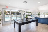 Located at 24432 Valencia Way in Laguna Niguel, California, this single-story clubhouse offers a welcoming, coastal-contemporary atmosphere designed for community gathering and relaxation. The open-plan layout is anchored by a spacious lounge area with soaring vaulted ceilings that amplify the sense of openness and allow natural light to pour throughout the space. Large floor-to-ceiling sliding glass doors line the exterior wall, providing seamless visual and physical access to the outdoor pool deck and patio area beyond, where lounge chairs and umbrella-shaded seating are visible.

The lounge is furnished with plush, curved navy-blue sectional seating and complementary aqua accent chairs, arranged around a low round coffee table — all accented by lush wall-mounted planters and potted greenery that reinforce the fresh, coastal aesthetic. A wall-mounted flat-screen television and a textured accent wall add functional and visual interest to the seating area.

The adjacent kitchen is equally impressive, featuring vaulted ceilings, warm wood-tone cabinetry, stainless steel appliances, a glass-top center island with bar seating, and a striking sculptural pendant light fixture. Decorative surfboard art mounted high on the kitchen walls adds a playful nod to the Southern California surf culture. A gallery hallway displaying coastal-themed artwork connects to additional areas of the clubhouse.