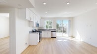 This single-floor unit showcases a clean, contemporary design characterized by crisp white walls, light hardwood flooring, and recessed lighting throughout. The open-concept kitchen serves as a central feature, equipped with white shaker-style cabinetry, gray countertops, and a full suite of stainless steel appliances including a gas range, microwave, and dishwasher. Large windows and a sliding glass door flood the kitchen with natural light while offering views of surrounding trees and outdoor scenery, with direct access to a balcony or outdoor area.

The bedroom is a bright, well-proportioned space featuring a large window framing outdoor views of palm trees and open sky. Built-in closet storage with shelving and a hanging rod provides practical organization, complemented by a sliding door for a streamlined look.

The bathroom maintains the unit's consistent aesthetic with light hardwood flooring, white walls, built-in shelving, and a tub-shower combination enclosed by sliding glass doors with chrome hardware. The 3D floor plan reveals a functional, efficiently arranged layout that maximizes the available space, with rooms flowing logically from the kitchen and living area into the private sleeping and bathroom spaces.