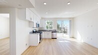 This single-floor unit showcases a clean, contemporary design characterized by crisp white walls, light hardwood flooring, and recessed lighting throughout. The open-concept kitchen serves as a central feature, equipped with white shaker-style cabinetry, gray countertops, and a full suite of stainless steel appliances including a gas range, microwave, and dishwasher. Large windows and a sliding glass door flood the kitchen with natural light while offering views of surrounding trees and outdoor scenery, with direct access to a balcony or outdoor area.

The bedroom is a bright, well-proportioned space featuring a large window framing outdoor views of palm trees and open sky. Built-in closet storage with shelving and a hanging rod provides practical organization, complemented by a sliding door for a streamlined look.

The bathroom maintains the unit's consistent aesthetic with light hardwood flooring, white walls, built-in shelving, and a tub-shower combination enclosed by sliding glass doors with chrome hardware. The 3D floor plan reveals a functional, efficiently arranged layout that maximizes the available space, with rooms flowing logically from the kitchen and living area into the private sleeping and bathroom spaces.