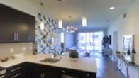 This single-floor space is defined by its open-concept layout, where the kitchen and living areas flow naturally into one another. The kitchen serves as a visual centerpiece, anchored by a spacious dark-cabinetry island with a built-in sink and light-colored countertops that provide strong contrast. Pendant lighting hangs above the island, complementing additional overhead fixtures that illuminate the space evenly.

The most distinctive architectural feature is the bold geometric accent wall, dressed in a blue-and-white patterned tile or wallcovering that adds a dynamic artistic focal point to the kitchen zone. A circular mirror mounted against this backdrop further enhances the visual depth of the area.

The living space beyond the kitchen benefits from abundant natural light streaming through large sliding glass doors, which appear to open onto an outdoor area, creating a seamless indoor-outdoor connection. A chandelier with an elegant branching design adds a decorative touch overhead. The overall ambiance is modern and stylish, balancing bold design choices with an open, airy feel reinforced by high ceilings and generous light throughout.