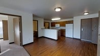 This single-floor unit is designed with an open-concept layout that seamlessly connects the living area to the kitchen. The flooring throughout the main space consists of warm-toned hardwood-style planks, which contrast with the light-colored walls and dark baseboards, creating a clean and cohesive aesthetic.

The kitchen is positioned at the rear of the open space and is equipped with dark wood cabinetry that provides ample storage. A peninsula-style counter extends outward, offering additional workspace and serving as a natural divider between the kitchen and living area. White appliances, including a refrigerator, microwave, and range, are integrated into the cabinetry layout. The countertops feature a stone-toned surface that complements the overall color palette.

Ceiling-mounted light fixtures provide consistent illumination throughout the space. A doorway visible from the main living area leads to what appears to be a bathroom, suggesting convenient access to additional amenities from the central living zone. The overall ambiance is practical and straightforward, suited for everyday living with a focus on functional use of space.