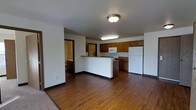 This single-floor unit is designed with an open-concept layout that seamlessly connects the living area to the kitchen. The flooring throughout the main space consists of warm-toned hardwood-style planks, which contrast with the light-colored walls and dark baseboards, creating a clean and cohesive aesthetic.

The kitchen is positioned at the rear of the open space and is equipped with dark wood cabinetry that provides ample storage. A peninsula-style counter extends outward, offering additional workspace and serving as a natural divider between the kitchen and living area. White appliances, including a refrigerator, microwave, and range, are integrated into the cabinetry layout. The countertops feature a stone-toned surface that complements the overall color palette.

Ceiling-mounted light fixtures provide consistent illumination throughout the space. A doorway visible from the main living area leads to what appears to be a bathroom, suggesting convenient access to additional amenities from the central living zone. The overall ambiance is practical and straightforward, suited for everyday living with a focus on functional use of space.
