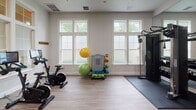 Located at 386 Attenborough Dr in Rosedale, MD, this single-floor fitness center is thoughtfully designed to support a comprehensive workout experience. The space is bright and open, with multiple large windows along the walls that invite an abundance of natural light and offer views of the surrounding greenery, creating a refreshing and motivating environment.

The cardio area is well-appointed with treadmills, elliptical trainers, and a stationary bike, all positioned beneath a wall-mounted flat-screen television for entertainment during workouts. A ceiling fan overhead ensures comfortable air circulation throughout the space.

The strength training section features a cable machine, a dumbbell rack with a full range of free weights, and an adjustable workout bench, complemented by floor-to-ceiling mirrors that enhance the sense of space and allow for form monitoring. Anti-fatigue rubber mats are placed beneath the equipment for safety and comfort.

A dedicated cycling area includes a Peloton-style interactive bike, and a separate accessory station offers stability balls and medicine balls for functional fitness. The light wood-toned flooring runs throughout, lending a clean, modern aesthetic to the entire facility. A set of glass-panel French doors connects to an adjoining room, adding to the facility's versatility.