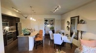 This single-story space in Midland presents an open-concept layout that seamlessly connects the kitchen and dining areas, creating a cohesive and functional living environment. The kitchen features a granite-topped island with bar seating accommodated by warm cognac-colored bar stools, illuminated by a trio of pendant lights that add a refined touch to the space. Dark hardwood flooring runs throughout, grounding the interior with a rich, elegant aesthetic that contrasts beautifully with the crisp white walls and cabinetry.

The dining area sits adjacent to the kitchen and is anchored by a round dining table surrounded by upholstered white chairs with nailhead trim detailing, lending a classic and polished feel. A decorative runner and centerpiece adorn the table, adding warmth to the setting. Track lighting overhead ensures the space is well-lit and practical.

A built-in shelving unit positioned between doorways provides both storage and display opportunities, adding character and functionality to the shared living space. Decorative wall accents and a large ornate mirror enhance the overall ambiance, making this space feel curated and welcoming throughout.