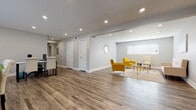 This single-floor commercial space offers a well-organized, modern environment designed to support a variety of professional functions. Throughout the entire space, warm hardwood-style flooring, soft gray walls, recessed lighting, and white baseboards create a clean, cohesive aesthetic.

The open-plan work area provides generous room for multiple workstations, with cream upholstered office chairs arranged around sleek black desks. Natural light filters in through several windows fitted with white horizontal blinds, keeping the atmosphere bright and inviting.

A welcoming lounge area is furnished with bold yellow accent chairs, a tufted cream sofa, and a round glass-and-gold coffee table atop a neutral area rug. A circular mirror on the wall and abstract wall art add refined decorative touches. This area transitions seamlessly into a smaller, more private seating nook well-suited for one-on-one meetings or consultations.

A dedicated meeting room accommodates a rectangular wood-and-metal dining-style table with seating for five, offering a focused setting for group discussions.

The kitchen is fully equipped with white shaker cabinetry, granite-look countertops, stainless steel appliances including a range, dishwasher, refrigerator, and microwave, providing excellent day-to-day functionality. An adjacent open shelving area offers additional storage and connects back to the main workspace.