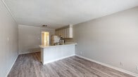 This single-floor unit is designed with a practical, open layout that connects the kitchen and living area into one cohesive space. The kitchen features a peninsula-style breakfast bar that separates it from the living area while maintaining an open feel. Upper and lower cabinetry in a light neutral finish provides ample storage, and the kitchen is equipped with a coil-burner electric range, range hood, and a refrigerator. A textured ceiling runs throughout the unit, and wood-style laminate flooring is consistent across the main living areas, adding warmth to the neutral gray-toned walls with white baseboards.

The living area is spacious and open, anchored by a sliding glass door fitted with vertical blinds that allow natural light to filter in while offering privacy and outdoor access. The layout, as seen in the 3D model, follows a linear floor plan with the kitchen and entry positioned at one end and the living space extending toward the sliding door.

The bathroom includes full square tile wainscoting along the walls, a tub-shower combination, a wall-mounted sink, toilet, and an overhead storage cabinet, offering functional use of the space. Overall, the unit presents a clean, straightforward interior with practical finishes suited for everyday living.