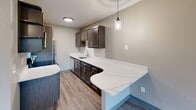 This single-floor space offers a well-organized layout with thoughtfully updated interiors. A central hallway connects the main living areas, finished with rich, dark hardwood-style flooring that runs consistently throughout. The living area receives generous natural light through large windows overlooking lush, tree-lined surroundings, and features a textured ceiling and a decorative chandelier that adds a refined touch. Baseboard heating units are present along the exterior walls.

The kitchen is a standout feature, showcasing dark espresso cabinetry, a subway tile backsplash, and an expansive quartz countertop with subtle veining that extends into a curved peninsula — ideal for additional workspace or casual dining. Pendant lighting and a stainless steel sink complement the design, while a full-size refrigerator completes the space.

The bedroom areas are spacious and bright, with the same hardwood-style flooring, neutral taupe walls, and windows that bring in natural light. The bathroom includes a full soaking tub and shower combination, finished with large-format light stone-effect tile and oil-rubbed bronze fixtures, offering a clean and contemporary aesthetic throughout.