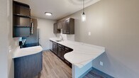 This single-floor space offers a well-organized layout with thoughtfully updated interiors. A central hallway connects the main living areas, finished with rich, dark hardwood-style flooring that runs consistently throughout. The living area receives generous natural light through large windows overlooking lush, tree-lined surroundings, and features a textured ceiling and a decorative chandelier that adds a refined touch. Baseboard heating units are present along the exterior walls.

The kitchen is a standout feature, showcasing dark espresso cabinetry, a subway tile backsplash, and an expansive quartz countertop with subtle veining that extends into a curved peninsula — ideal for additional workspace or casual dining. Pendant lighting and a stainless steel sink complement the design, while a full-size refrigerator completes the space.

The bedroom areas are spacious and bright, with the same hardwood-style flooring, neutral taupe walls, and windows that bring in natural light. The bathroom includes a full soaking tub and shower combination, finished with large-format light stone-effect tile and oil-rubbed bronze fixtures, offering a clean and contemporary aesthetic throughout.
