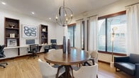 Located in Bella Terra Center in Richmond, TX, this single-floor office space is thoughtfully designed to support both focused work and collaborative meetings. The room is finished with warm light hardwood flooring throughout, complemented by crisp white walls and rich dark wood trim that frames the windows and built-in shelving, lending the space a polished, professional aesthetic.

A central round wooden conference table surrounded by upholstered chairs serves as the meeting focal point, anchored above by a striking spherical chandelier that adds a touch of contemporary elegance. Recessed lighting further enhances the ambient illumination. Along one wall, a built-in desk with dual workstations is flanked by open wooden bookcases, providing ample workspace and storage for office essentials.

Multiple large windows dressed with sheer white curtains and adjustable blinds invite generous natural light while offering exterior views of the surrounding greenery. A wall-mounted flat-screen TV and a decorative console table round out the room's functionality. A leather accent chair and curated artwork complete the refined, modern-professional atmosphere, making this office equally suitable as a productive workspace and an inviting meeting environment.