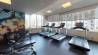 This fitness center is a comprehensively equipped space designed to support a full range of workout routines. The layout is thoughtfully organized, with distinct zones dedicated to cardio, strength training, and free weights. Floor-to-ceiling windows line the perimeter, fitted with adjustable roller shades that allow natural light to fill the space while framing sweeping views of the surrounding urban skyline — a standout feature that enhances the workout experience.

The cardio section is outfitted with multiple treadmills, elliptical machines, stair climbers, and stationary cycling bikes, all positioned to take advantage of the city views. Wall-mounted flat-screen televisions provide additional entertainment options throughout this zone.

The strength training area includes multi-functional cable machines and a well-stocked free weight rack with a full range of dumbbells, accompanied by adjustable benches. Full-length mirrors along one wall expand the sense of space and aid in proper form during exercise.

Durable rubber flooring covers the entire space, and recessed panel lighting in the ceiling ensures consistent illumination. A vibrant, abstract mural adds a dynamic artistic element, contributing to an energetic and modern ambiance throughout.