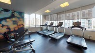 This fitness center is a comprehensively equipped space designed to support a full range of workout routines. The layout is thoughtfully organized, with distinct zones dedicated to cardio, strength training, and free weights. Floor-to-ceiling windows line the perimeter, fitted with adjustable roller shades that allow natural light to fill the space while framing sweeping views of the surrounding urban skyline — a standout feature that enhances the workout experience.

The cardio section is outfitted with multiple treadmills, elliptical machines, stair climbers, and stationary cycling bikes, all positioned to take advantage of the city views. Wall-mounted flat-screen televisions provide additional entertainment options throughout this zone.

The strength training area includes multi-functional cable machines and a well-stocked free weight rack with a full range of dumbbells, accompanied by adjustable benches. Full-length mirrors along one wall expand the sense of space and aid in proper form during exercise.

Durable rubber flooring covers the entire space, and recessed panel lighting in the ceiling ensures consistent illumination. A vibrant, abstract mural adds a dynamic artistic element, contributing to an energetic and modern ambiance throughout.