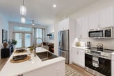 Located at 333 W 24th St in Houston's vibrant Heights neighborhood, this one-bedroom, one-bathroom residence offers a thoughtfully designed open-concept layout finished with contemporary details. The kitchen is a standout feature, boasting crisp white shaker cabinetry extending to the ceiling, a tile backsplash, stainless steel appliances, quartz countertops, and a central island with bar seating — all illuminated by elegant pendant lighting. Light wood-tone flooring flows seamlessly into the adjoining living room, where large windows and glass French doors invite abundant natural light and connect the interior to the outdoors. A ceiling fan adds comfort to the living area. The bedroom is a calm retreat, featuring a generous window with patterned drapes, wood-style flooring, and ample space for a sizable bed and storage. The bathroom impresses with marble-look tile flooring, a dual-sink white vanity, a walk-in marble-clad shower, and direct access to a dedicated closet. Throughout, the design balances modern aesthetics with functional living, creating a cohesive, comfortable space well-suited for everyday life in one of Houston's most desirable urban neighborhoods.