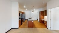 This single-floor residence offers a clean, modern layout designed for comfortable everyday living. The heart of the home is an open-concept kitchen and living area, where warm medium-toned wood cabinetry pairs with light countertops and a full suite of stainless steel appliances, including a refrigerator, electric range, and over-the-range microwave. A central island with an undermount sink serves as both a functional workspace and a natural divider between the kitchen and the living area, accented above by stylish pendant lighting. The kitchen also features tile backsplash and ample upper and lower cabinet storage. Hardwood-style flooring runs through the kitchen and entry areas, while plush carpet transitions into the living and bedroom spaces, adding warmth underfoot. The bedroom is a bright, comfortably sized room with a window that allows natural light to fill the space. The bathroom is clean and functional, featuring a walk-in shower, a vanity with sink, and tile flooring. A dedicated in-unit laundry room houses a full-sized side-by-side washer and dryer with wire shelving above for additional storage. Recessed lighting and large windows throughout keep the space feeling bright and open.