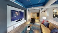 This stylish amenity lounge is defined by its bold, contemporary design — most notably a striking teal-painted ceiling with exposed ductwork and piping that adds an industrial-chic character throughout the space. Light wood-plank flooring flows seamlessly across the open layout, tying together distinct activity zones with a cohesive aesthetic.

At the heart of the space is a sleek, modern billiard table with a white base and teal felt surface, positioned beneath a sculptural branching chandelier. Large floor-to-ceiling windows dressed in bold teal-and-white striped curtains flood the room with natural light and offer views of the surrounding outdoors.

A dedicated entertainment lounge area features a large wall-mounted flat-screen TV paired with built-in white cabinetry, a glass coffee table, and comfortable seating. An oversized Scrabble board wall display adds a playful, social element to the décor.

The kitchenette area is outfitted with a wall oven, refrigerator, and dark countertops set against a shimmering teal mosaic tile backsplash, providing functional support for gatherings. Decorative art panels featuring fluid teal-and-white abstract designs are integrated into the walls, reinforcing the space's vibrant color palette. Overall, the lounge delivers a dynamic, socially engaging environment with thoughtful design details throughout.
