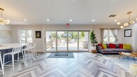 Located at 3801 Memory Lane in Abingdon, Maryland, this community clubhouse is a thoughtfully designed single-floor gathering space that blends modern aesthetics with welcoming functionality. The standout herringbone-patterned luxury vinyl flooring flows seamlessly throughout the entire space, tying together its distinct zones with visual continuity.

The main lounge area is bright and inviting, anchored by a bold teal accent wall and illuminated by a striking mid-century modern globe chandelier. Colorful, contemporary furnishings — including a grey sofa, vibrant yellow armchairs, and layered rugs — create a relaxed and lively atmosphere. A wall-mounted flat-screen TV adds to the space's entertainment appeal, while multiple windows flood the room with natural light.

Large sliding glass doors open directly to an outdoor patio with stone pillars and seating areas framed by lush greenery, seamlessly connecting indoor and outdoor living. A welcoming entry mat greets visitors at the threshold.

The fully equipped kitchen features rich dark wood cabinetry, a vivid green subway tile backsplash, white quartz countertops, stainless steel appliances, and a spacious center island with bar seating — ideal for community gatherings and events. Recessed lighting and a window overlooking the outdoors keep the kitchen bright and functional. Together, these elements make this clubhouse a well-appointed and energetic hub for community life.