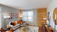 This single-floor residence is thoughtfully designed with a cohesive modern aesthetic and warm, earthy tones throughout. The living area serves as the heart of the home, featuring rich hardwood flooring, soft gray walls, and a striking natural wood plank accent wall that adds warmth and texture to the space. A large window dressed with geometric-patterned curtains fills the room with natural light and offers views of the surrounding exterior. A dedicated workspace sits adjacent to the living area, benefiting from the same natural light and accent wall backdrop.

The kitchen is designed for both function and style, showcasing dark cabinetry paired with granite countertops and black stainless steel appliances, including a refrigerator, range, and built-in microwave. A peninsula with an extended granite overhang accommodates bar seating for four, illuminated by sleek pendant lighting — an ideal setup for casual dining or entertaining.

The bedroom features plush carpeting, neutral walls, and a well-sized window with patterned curtains that allow ample natural light. The bathroom includes a full soaking tub and shower combination with tile surround. A dedicated laundry area with in-unit washer provides added convenience, complemented by wire shelving for storage.