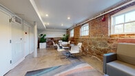 Located at 9 Mill St in Paterson, NJ, this single-floor commercial space masterfully combines historic architectural character with contemporary design. The interior is defined by striking exposed brick and original stone masonry walls, which serve as a compelling backdrop throughout the space. Natural light filters in through multiple double-hung windows set within the brick facade, illuminating the polished concrete flooring and creating a warm, inviting atmosphere.

The main area is organized into distinct functional zones. A collaborative seating area features dark wood tables paired with modern white perforated chairs, ideal for meetings or casual work sessions. A lounge zone offers a generously sized gray sectional sofa arranged around a wood-topped coffee table, anchored by a colorful, wave-patterned area rug that adds a vibrant contrast to the industrial surroundings. Large tropical potted plants placed throughout the space introduce organic warmth and visual interest.

White structural columns punctuate the interior, complementing the clean white walls and ceilings with recessed lighting. Exposed utility piping adds to the industrial aesthetic. Additional doors along one wall suggest access to private rooms or utilities, contributing to the overall functionality of this versatile, characterful space.