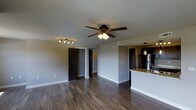 This single-floor space offers a well-designed, open-concept layout that seamlessly connects the living area to the kitchen. The living area is finished with rich, dark hardwood flooring that runs throughout, complemented by clean white baseboards and neutral gray walls that create a modern, cohesive aesthetic. A multi-blade ceiling fan with integrated lighting serves as a functional centerpiece overhead, while a track lighting fixture near the entryway adds additional illumination to the space.

A walk-in closet is accessible from the living area, offering generous storage behind a dark-paneled door. The open passageway leads further into the unit, suggesting a thoughtful, flowing floor plan.

The kitchen is defined by a peninsular breakfast bar with a granite or stone-finish countertop, providing both workspace and casual seating opportunities. Dark cabinetry lines the kitchen walls, offering ample storage, while stainless steel appliances — including a full-size refrigerator — add a sleek, contemporary touch. A pendant light fixture hangs above the kitchen area, enhancing its warm ambiance.

Overall, the space presents a clean, modern interior with consistent dark and neutral tones, combining practicality with style across its open, single-level layout.