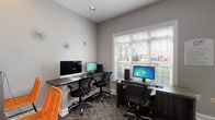 This single-room business center is thoughtfully designed to provide a fully functional workspace for residents or guests. The room features patterned carpet flooring, neutral gray walls, and clean white trim throughout, creating a professional yet approachable atmosphere.

Multiple dedicated workstations are arranged throughout the space, each equipped with desktop computers and ergonomic task chairs. A wall-mounted counter along one side provides additional workspace and houses a shared printer, making document management convenient. Vibrant orange accent chairs add a pop of color and offer supplementary seating at the counter.

Natural light enters through a large window fitted with horizontal blinds, brightening the workspace while allowing for adjustable light control. A French-style glass-paned door connects the business center to an adjacent common area, visible in the background and featuring its own seating, enhancing the sense of connectivity within the facility.

Organizational tools including a cork bulletin board display community notices and informational postings. Recessed ceiling lighting ensures consistent illumination throughout the room. The overall layout is efficient and purposeful, maximizing the available floor area to accommodate multiple users simultaneously, making it an ideal space for tasks such as printing, research, and remote work.