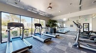 Located at 1100 E Bell Rd in Phoenix, AZ, this fitness center offers a comprehensive, single-floor exercise facility designed to support a wide range of workout routines. The space is thoughtfully laid out across a generously sized open floor plan, with patterned carpet tiles covering the entire area for comfort and durability.

The cardio zone features a row of treadmills and elliptical machines positioned along a floor-to-ceiling glass window wall, providing views of the surrounding outdoor landscape and abundant natural light. Ceiling fans and overhead fluorescent lighting ensure the space remains well-ventilated and brightly lit throughout.

The strength training section is well-appointed with a full rack of dumbbells, weight benches, and multiple cable and resistance machines. Large mirrors line the walls in this area, enhancing the sense of space while allowing users to monitor their form. An additional functional training zone includes TRX suspension straps, medicine balls, stability balls, a stationary bike, and a wall-mounted television for guided workouts.

A wooden storage unit provides organized access to exercise mats and accessories, while a water fountain is conveniently installed nearby. The facility is accessed through glass-paneled doors and is clearly marked with posted fitness center rules, reflecting an organized and user-focused environment.