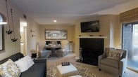 Located at 2225 Ashley River Road in Charleston, South Carolina, this single-floor residence offers a warm and inviting atmosphere with a thoughtfully designed interior. The living area serves as the heart of the home, centered around a striking fireplace with a bold dark surround and a clean white mantel, adding a classic focal point to the room. A wall-mounted flat-screen television above the mantel enhances the space's functionality without compromising its aesthetic appeal. Soft, neutral wall tones create a calming backdrop that complements the plush carpeted flooring, while a patterned area rug in muted grays and yellows adds a touch of visual interest underfoot. Comfortable seating, including upholstered armchairs and a sofa, invites relaxation throughout the space. A cozy dining nook features a round pedestal table accompanied by upholstered chairs, ideal for intimate gatherings. Coastal-inspired artwork, including a framed fish print, nods to the home's Charleston waterside setting and lends character to the walls. Recessed lighting provides a warm, ambient glow throughout. The overall ambiance is sophisticated yet approachable, blending casual comfort with refined Southern charm.